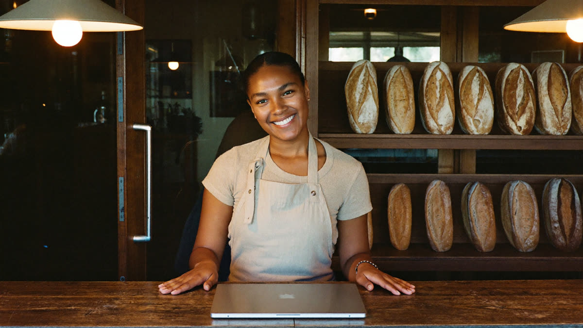 A smiling bakery owner stands at the counter at closing time next to a closed laptop