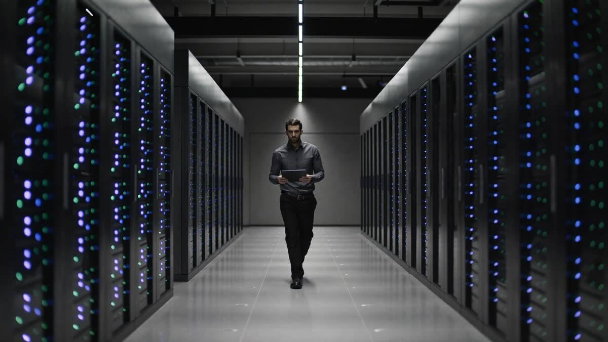 An engineer with a tablet walks down a long data centre aisle between server racks